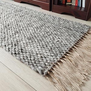 May include: A gray and white textured runner rug with a fringe detail. The rug is made of a thick, woven material and has a neutral color palette. The fringe is a light beige color.
