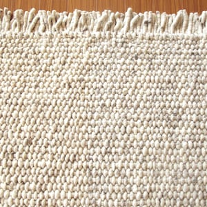 Handwoven chunky wool rug in natural undyed cream/ off-white and soft grey tones, shown in close-up with thick looped texture and fringe detail.