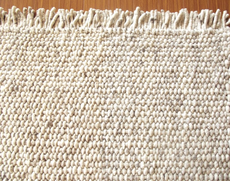 Handwoven chunky wool rug in natural undyed cream/ off-white and soft grey tones, shown in close-up with thick looped texture and fringe detail.