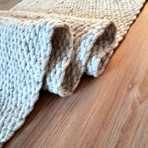 May include: A light gray runner rug featuring a thick, chunky knit texture with a repeating woven pattern. The rug is partially folded over, revealing the dense, looped construction of the yarn and the soft, neutral color of the material.