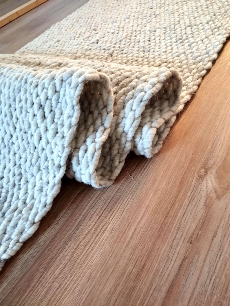 May include: A light gray runner rug featuring a thick, chunky knit texture with a repeating woven pattern. The rug is partially folded over, revealing the dense, looped construction of the yarn and the soft, neutral color of the material.