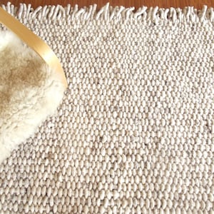 Top view of a handwoven chunky wool rug with thick looped texture and fringe, in natural off-white tones and grey speckled, partially shown with a light lounge chair in the corner. The rug is handmade of natural, undyed and untreated wool and has a minimalist, nordic, scandinavian style, suitable for any home decor.