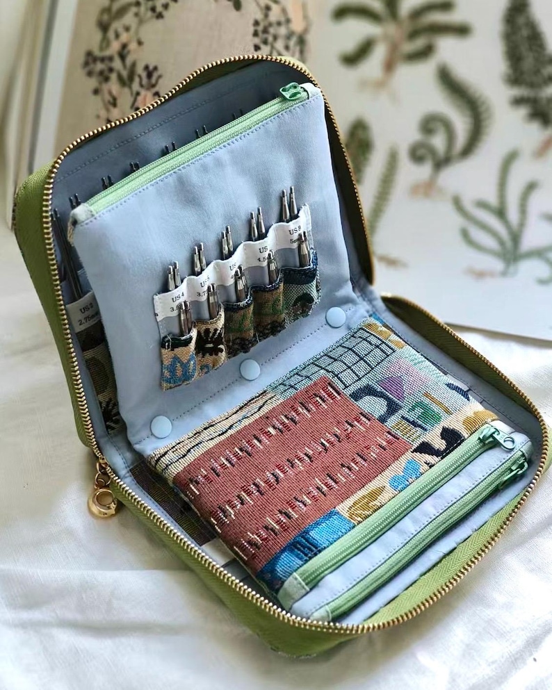 K&ouml;nnte beinhalten: Ein salbeigr&uuml;nes Etui mit Rei&szlig;verschluss und hellblauem Innenorganizer. Der Organizer verf&uuml;gt &uuml;ber mehrere F&auml;cher f&uuml;r Stricknadelspitzen aus Metall, eine gemusterte Stofftasche darunter und ein separates Rei&szlig;verschlussfach.