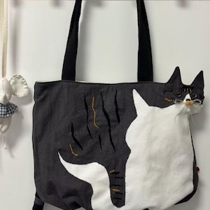 May include: A black and white cat-themed tote bag with black handles. The bag features a large white cat design on the front and a striped tail. The cat's head is on the side of the bag. The bag is hanging on a wooden rack.