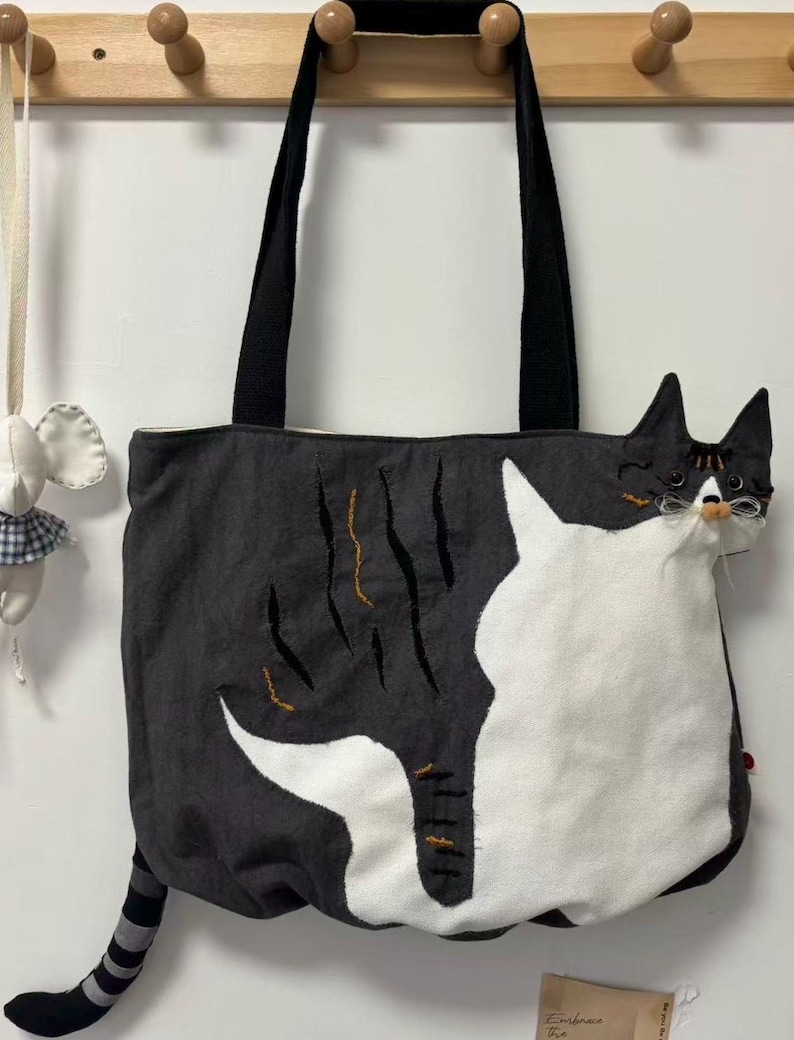 May include: A black and white cat-themed tote bag with black handles. The bag features a large white cat design on the front and a striped tail. The cat's head is on the side of the bag. The bag is hanging on a wooden rack.