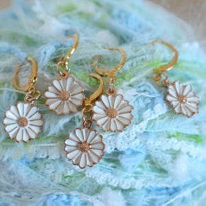 Può includere: Orecchini a margherita color oro con petali in smalto bianco e centro dorato. Gli orecchini sono su uno sfondo strutturato blu e verde. Hanno una chiusura a cerchio.