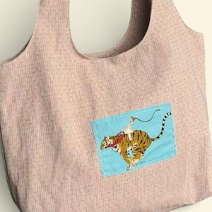May include: A light pink tote bag with a repeating vertical dotted stripe pattern. A blue square patch features a cartoon image of a person riding a tiger. The tiger is orange and black.
