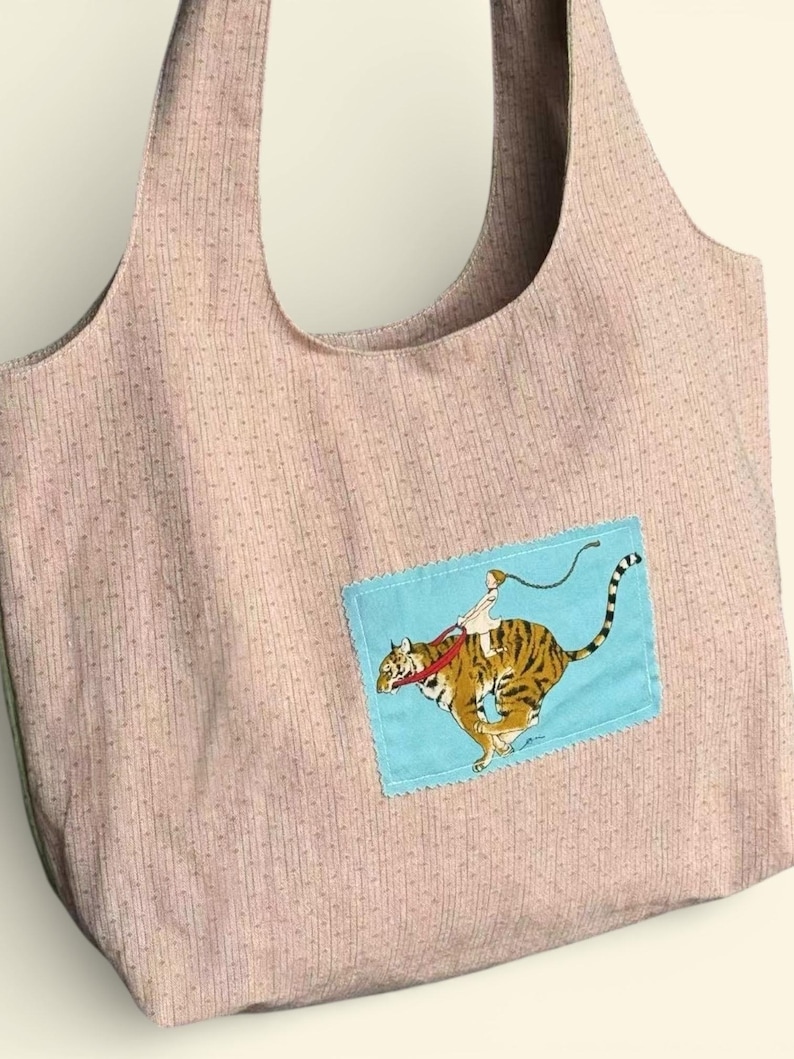 May include: A light pink tote bag with a repeating vertical dotted stripe pattern. A blue square patch features a cartoon image of a person riding a tiger. The tiger is orange and black.