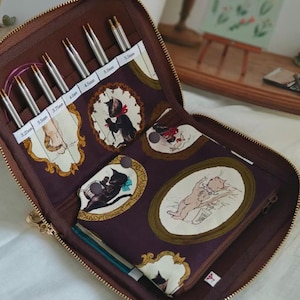Peut inclure: Une trousse à aiguilles à tricoter marron zippée avec un intérieur à motifs d'illustrations de chats. L'étui contient plusieurs aiguilles à tricoter argentées de différentes tailles, de 3,25 mm à 5,5 mm. Une petite pochette zippée est également visible.