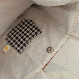 May include: Interior view of a beige tote bag with a brown leather trim. A small, brown and white checkered patch with a yellow heart tag is attached to the inside. A silver magnetic snap closure is visible.