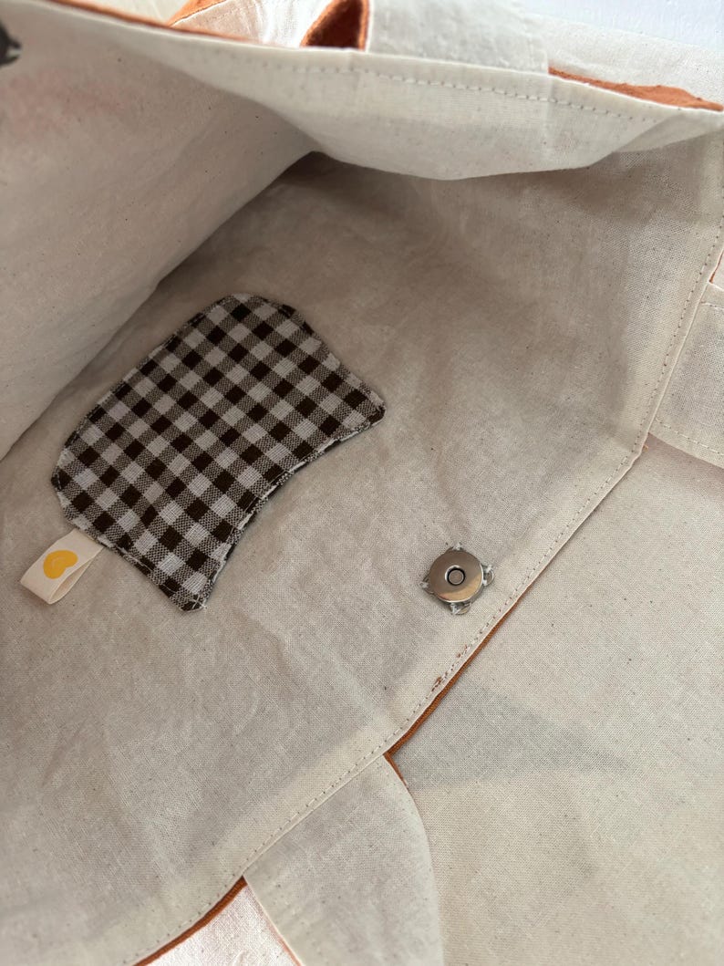May include: Interior view of a beige tote bag with a brown leather trim. A small, brown and white checkered patch with a yellow heart tag is attached to the inside. A silver magnetic snap closure is visible.