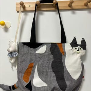 May include: A gray tote bag with a cat design. The bag features a black top and handles, with a patchwork design of gray, white, black, and orange shapes. The cat's face and tail are prominent, adding a whimsical touch. A small elephant-shaped ornament hangs nearby.
