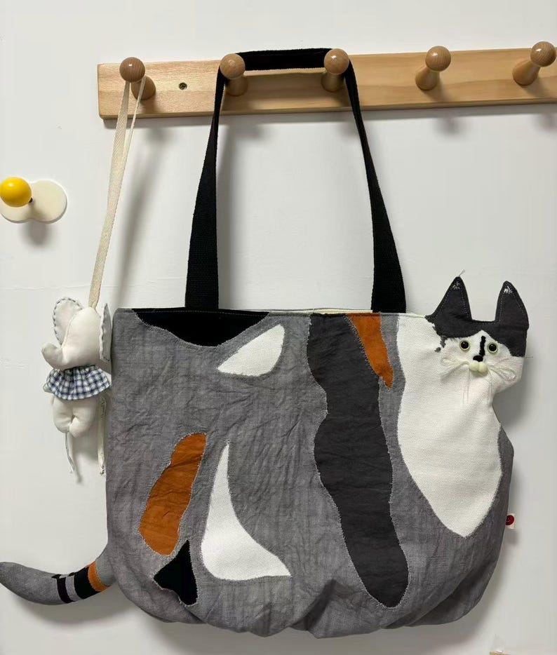 May include: A gray tote bag with a cat design. The bag features a black top and handles, with a patchwork design of gray, white, black, and orange shapes. The cat's face and tail are prominent, adding a whimsical touch. A small elephant-shaped ornament hangs nearby.