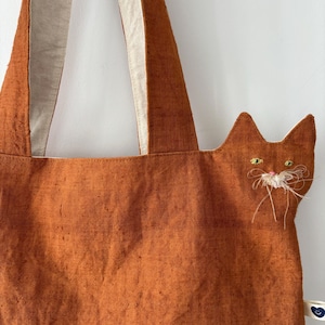 Op de afbeelding: Oranje draagtas met een kattengezichtontwerp. De tas heeft een textuurstof en cr&egrave;mekleurige bandjes. Het kattengezicht is geborduurd met gele ogen en witte snorharen. Een klein label met een blauw hart is aan de zijkant bevestigd.