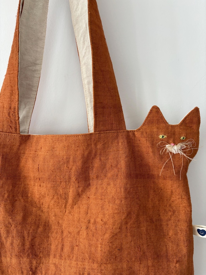 Op de afbeelding: Oranje draagtas met een kattengezichtontwerp. De tas heeft een textuurstof en cr&egrave;mekleurige bandjes. Het kattengezicht is geborduurd met gele ogen en witte snorharen. Een klein label met een blauw hart is aan de zijkant bevestigd.