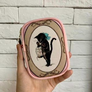 Op de afbeelding: Een roze en beige etui met ritssluiting met een illustratie van een zwarte kat met een blauwe strik die een vogelkooi vasthoudt. De ovale afbeelding is omlijst met een beige rand en bloemaccenten. De etui heeft een roze rand en een gouden rits.