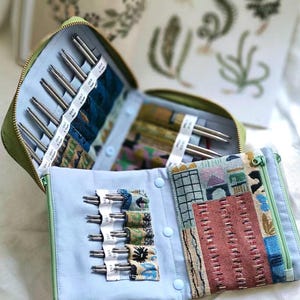 K&ouml;nnte beinhalten: Ein gr&uuml;nes Etui mit Rei&szlig;verschluss enth&auml;lt ein Set aus Metallstricknadeln, die in hellblauen Stofff&auml;chern organisiert sind. Das Innere verf&uuml;gt &uuml;ber gemusterte Stofftaschen und ein Rei&szlig;verschlussfach mit einem geometrischen und architektonischen Druck.