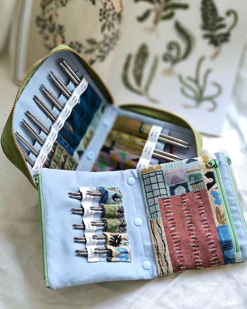 K&ouml;nnte beinhalten: Ein gr&uuml;nes Etui mit Rei&szlig;verschluss enth&auml;lt ein Set aus Metallstricknadeln, die in hellblauen Stofff&auml;chern organisiert sind. Das Innere verf&uuml;gt &uuml;ber gemusterte Stofftaschen und ein Rei&szlig;verschlussfach mit einem geometrischen und architektonischen Druck.