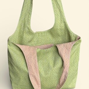 May include: A green tote bag with a textured pattern and pink straps. The bag is open, revealing a pink interior. The straps are wide and appear to be made of the same material as the interior. The bag is designed for carrying items.
