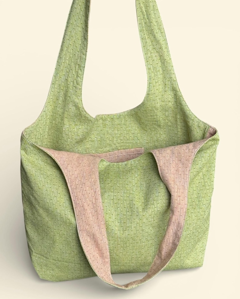 May include: A green tote bag with a textured pattern and pink straps. The bag is open, revealing a pink interior. The straps are wide and appear to be made of the same material as the interior. The bag is designed for carrying items.
