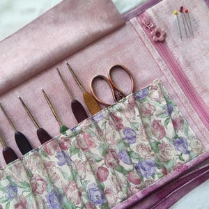 Peut inclure: Une trousse à crochets rose avec une poche à imprimé floral. La trousse contient plusieurs crochets avec des manches en bois et colorés, une paire de ciseaux en cuivre et quelques épingles. La fermeture éclair a une tirette en forme de fleur.