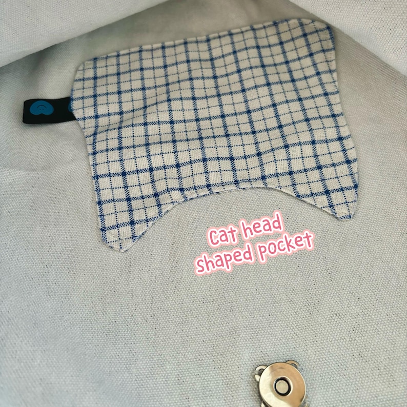 May include: A cat head-shaped pocket made of fabric with a blue and white plaid pattern. The pocket has a black tab with a blue heart-shaped detail. The text "Cat head shaped pocket" is visible.