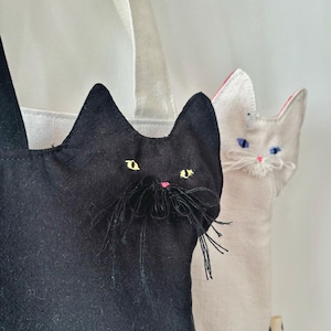 May include: Two cat-shaped tote bags. One is black with yellow eyes and a pink nose, the other is off-white with blue eyes and a pink nose. Both have long black whiskers and are made of fabric with white handles.