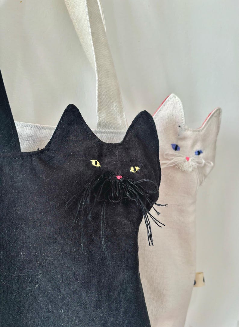 May include: Two cat-shaped tote bags. One is black with yellow eyes and a pink nose, the other is off-white with blue eyes and a pink nose. Both have long black whiskers and are made of fabric with white handles.