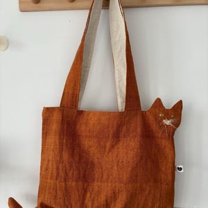 Op de afbeelding: Oranje tote bag met een kattenontwerp. De tas heeft een kattengezicht met geborduurde ogen en snorharen, en een staart. De handvatten zijn aan de binnenkant lichter van kleur. Een charmant en uniek accessoire.