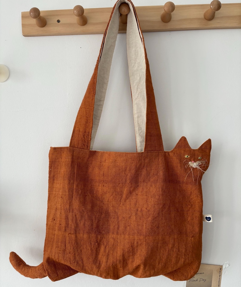 Op de afbeelding: Oranje tote bag met een kattenontwerp. De tas heeft een kattengezicht met geborduurde ogen en snorharen, en een staart. De handvatten zijn aan de binnenkant lichter van kleur. Een charmant en uniek accessoire.