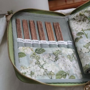 Può includere: Un astuccio aperto con cerniera verde oliva con interno floreale, contenente diversi ferri da maglia in legno di varie dimensioni. Gli aghi sono etichettati con le loro dimensioni in millimetri, da 2,25 mm a 3,75 mm. L'astuccio ha una cerniera dorata.