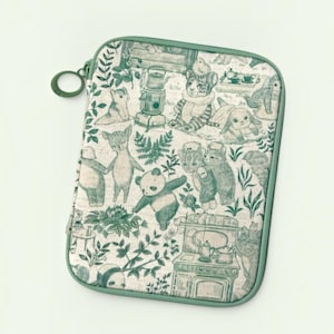 Puede incluir: Un estuche rectangular con cremallera que presenta una base de tela beige claro con un patrón de ilustraciones repetitivas en verde oscuro. El diseño muestra varios animales, incluyendo un panda, un tigre y conejos, junto a objetos domésticos como una estufa.