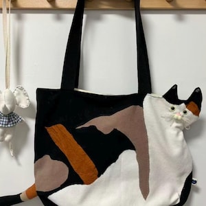 May include: Black tote bag with a cat design. The bag features a black background with patches of brown, orange, and white fabric. A three-dimensional cat head and tail are attached to the bag. The bag has black handles.