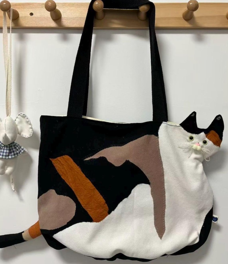 May include: Black tote bag with a cat design. The bag features a black background with patches of brown, orange, and white fabric. A three-dimensional cat head and tail are attached to the bag. The bag has black handles.