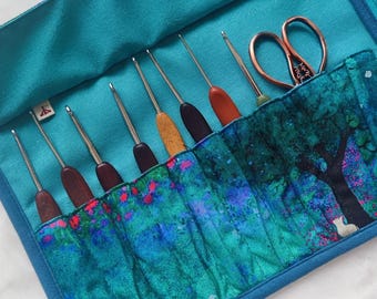 Puede incluir: Un estuche turquesa para ganchillos con un diseño de árbol colorido. El estuche contiene varios ganchillos con mangos de madera y marrón oscuro, además de unas tijeras de cobre. El estuche está abierto, mostrando las herramientas.