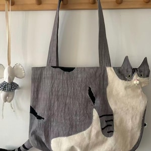 May include: Gray tote bag with a cat design. The bag features a gray body with black accents and a white and gray cat face on the side. The cat's tail extends from the bottom of the bag. The bag has two gray handles.