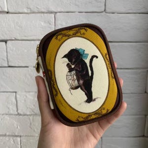 Può includere: Un piccolo portamonete rettangolare con un motivo decorativo a gatto. La borsa presenta un gatto nero con un fiocco blu che tiene una gabbia per uccelli, su uno sfondo color crema all'interno di una cornice ovale dorata. La borsa ha un bordo marrone e una cerniera dorata.