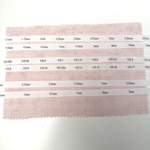 May include: White ribbons with printed measurements in millimeters and US sizes are displayed on a pink fabric swatch. The ribbons show sizes from 1.5mm to 12mm and US sizes from 000 to 17. The image is taken from above.
