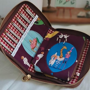 Puede incluir: Estuche de agujas de tejer marrón abierto con forro a cuadros rojos y blancos. El estuche contiene varias agujas de tejer plateadas de varios tamaños y una tela estampada con ilustraciones caprichosas.