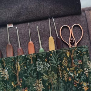 May include: A dark brown fabric roll-up organizer case featuring a green corduroy pocket with a forest-themed print of trees, flowers, and small animals. The case holds six metal crochet hooks with various wooden handles and a pair of copper scissors.