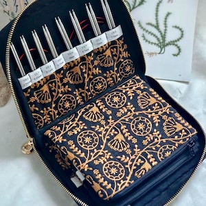 May include: A black zipped case containing a set of interchangeable knitting needles. The interior features gold floral embroidery on black fabric. The needles are labelled with sizes US 2 (2.75mm) through US 9 (5.5mm) and include attached red cables.