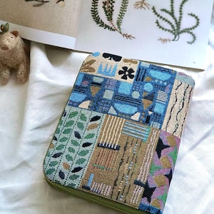 K&ouml;nnte beinhalten: Ein rechteckiges Etui mit Rei&szlig;verschluss aus gewebtem Stoff im Wandteppich-Stil mit einem mehrfarbigen geometrischen und botanischen Muster in Blau-, Gr&uuml;n-, Gold- und Schwarzt&ouml;nen sowie einem olivgr&uuml;nen Rei&szlig;verschlussrand.
