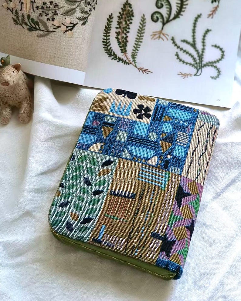 K&ouml;nnte beinhalten: Ein rechteckiges Etui mit Rei&szlig;verschluss aus gewebtem Stoff im Wandteppich-Stil mit einem mehrfarbigen geometrischen und botanischen Muster in Blau-, Gr&uuml;n-, Gold- und Schwarzt&ouml;nen sowie einem olivgr&uuml;nen Rei&szlig;verschlussrand.
