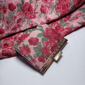 Puede incluir: Una tela rosa con estampado floral de rosas rojas y hojas verdes. Un pequeño bolso de mano cuadrado con un cierre dorado. La tela está doblada, mostrando el patrón floral repetido.