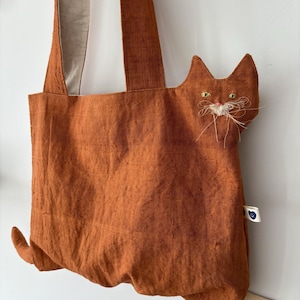 Op de afbeelding: Oranje draagtas met een kattenontwerp. De tas heeft een kattengezicht met geborduurde ogen en snorharen. De tas heeft lange banden en een staartdetail. De tas is gemaakt van een textuurstof.