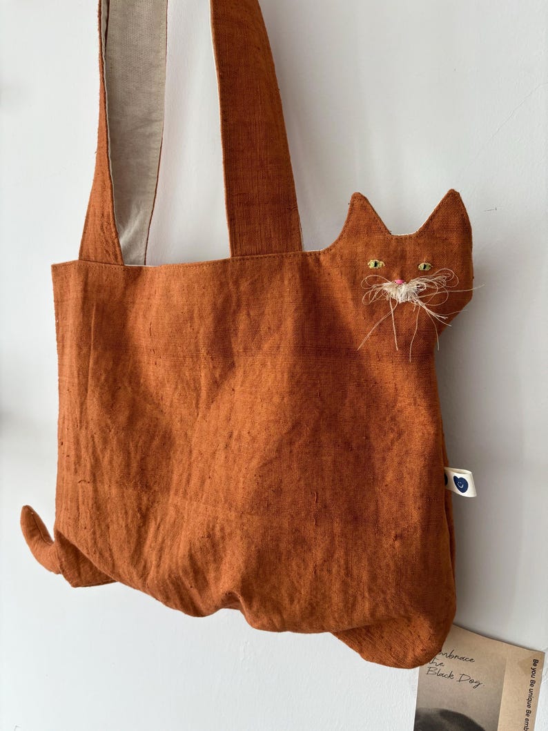 Op de afbeelding: Oranje draagtas met een kattenontwerp. De tas heeft een kattengezicht met geborduurde ogen en snorharen. De tas heeft lange banden en een staartdetail. De tas is gemaakt van een textuurstof.