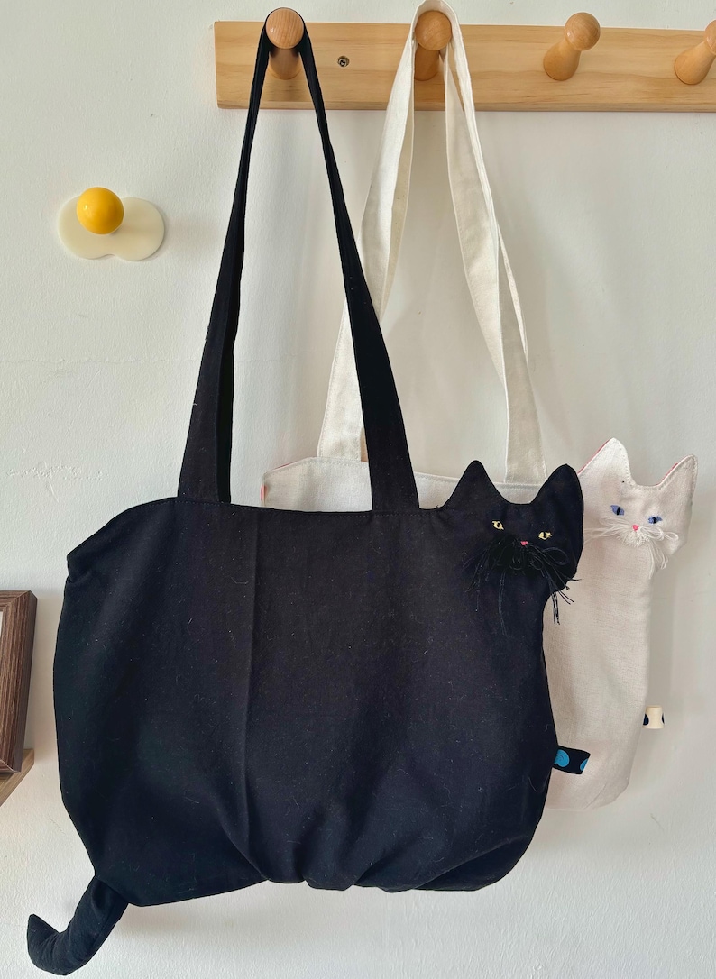 May include: Black tote bag with cat face detail, hanging next to a white cat-shaped tote bag. The black bag has a long tail and black handles. The white bag has blue eyes and pink nose. Both bags are hanging on a wooden rack.