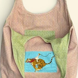 May include: A pink and green shoulder bag with a whimsical tiger design. The bag features a light pink fabric with a dotted pattern, green straps and a blue patch with a tiger and rider illustration.