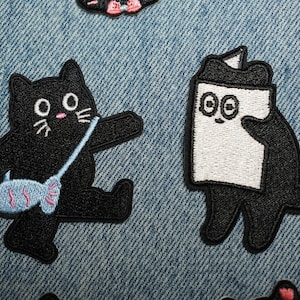 以下が含まれることがあります： 漫画の猫の刺繍パッチ。青いバッグと魚のデザインの黒猫1匹、白黒の猫のようなキャラクターがもう1匹。パッチは青いデニムの背景にあります。