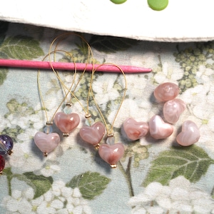 Könnte beinhalten: Nahaufnahme von herzförmigen Maschenmarkierern und losen Perlen. Die Maschenmarkierer haben rosa herzförmige Perlen, Golddraht und kleine blaue Perlen. Mehrere lose rosa herzförmige Perlen sind in der Nähe verstreut.